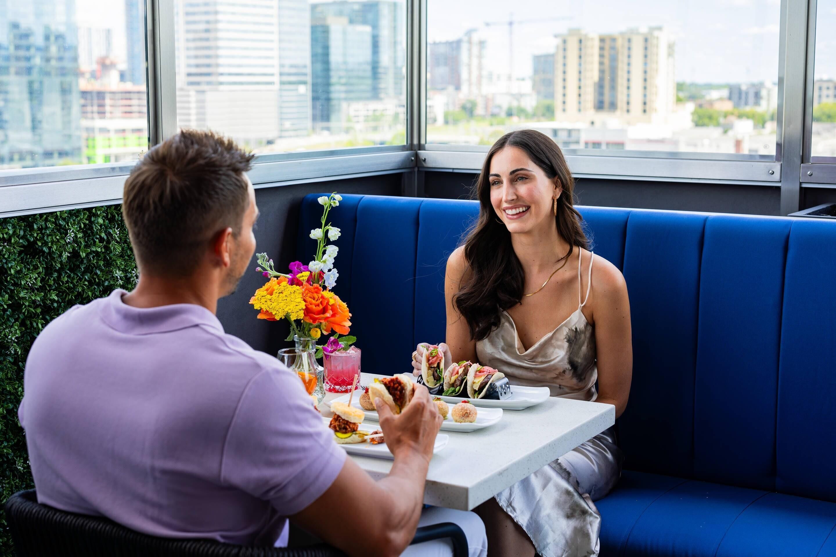 A couple dining at a table with a city view, a blue cushioned bench, and a floral centerpiece.