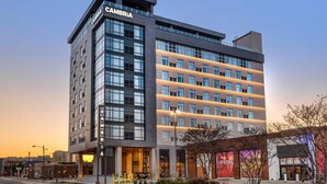 A modern hotel with a prominent "CAMBRIA" sign, featuring a mix of glass and concrete architecture, and a visible entrance with a parking area in front.