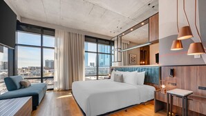 A modern hotel room with a large bed, a sofa, and a desk. The room has large windows offering a city view.