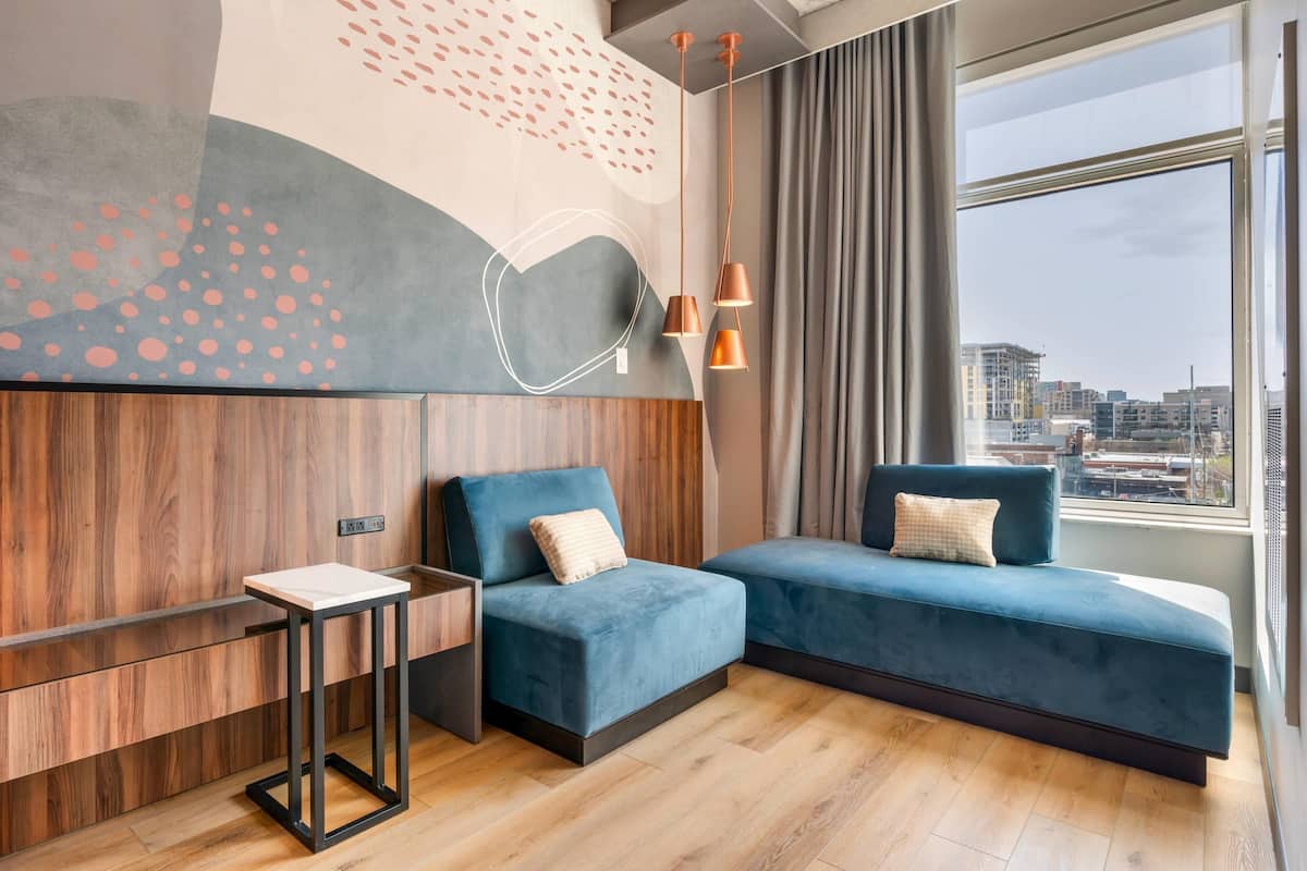 A modern hotel room with a large window, a blue sofa, a small table, and a unique wall design.