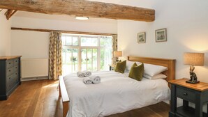 Ein Schlafzimmer mit Holzbalkendecke, einem großen Fenster mit Vorhängen, einem Bett mit weißen Leinen, zwei Nachttischlampen und einem kleinen Tisch mit Lampe.