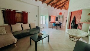A living room with a sofa, a coffee table, and a wicker chair. There is a dining area with a table and chairs in the background.