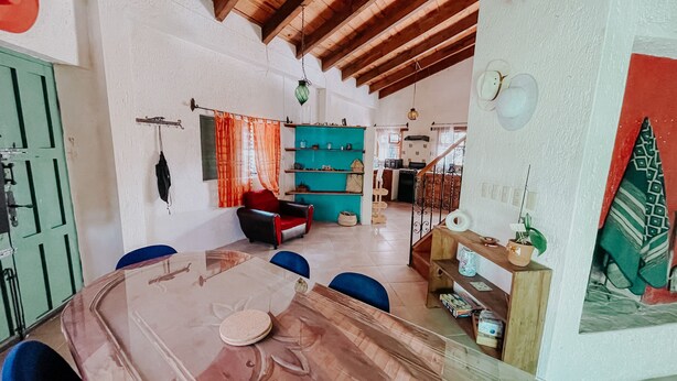 A dining area with a wooden table, chairs, and a red chair. There is a kitchen area with shelves and a hanging light.