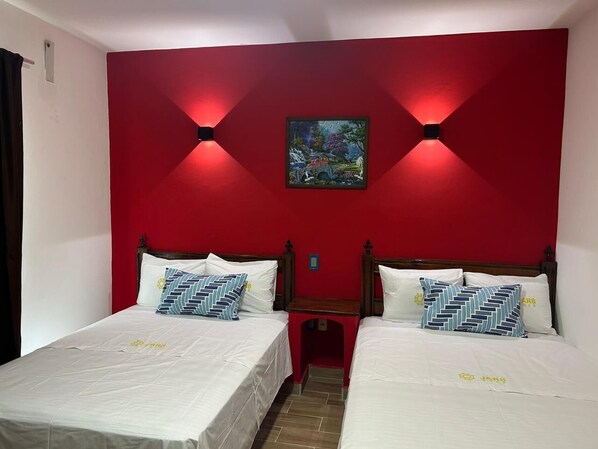 Habitación con dos camas, cada una con ropa de cama blanca y almohadas estampadas, frente a una pared roja con un cuadro enmarcado.