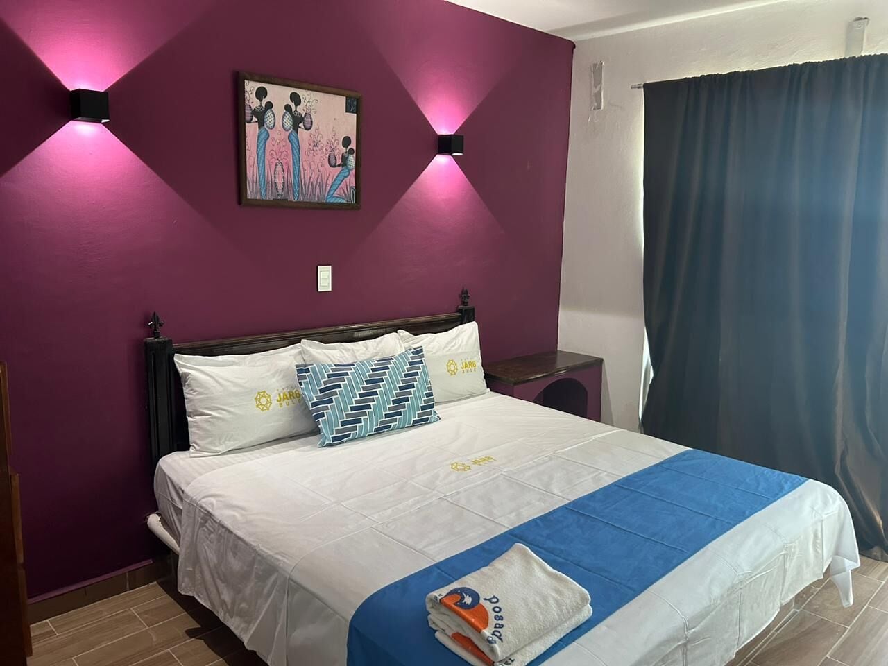 A bed with white bedding and a blue accent, a wall-mounted painting, and a window with a blue curtain.