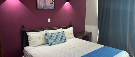 A bed with white bedding and a blue accent, a wall-mounted painting, and a window with a blue curtain.