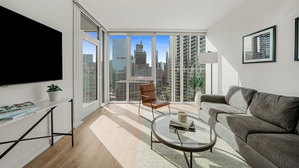A modern living room with a large TV, a sofa, a coffee table, and a chair. There is a large window with a city view. - Level River North - Spacious Executive Suite (Chicago)