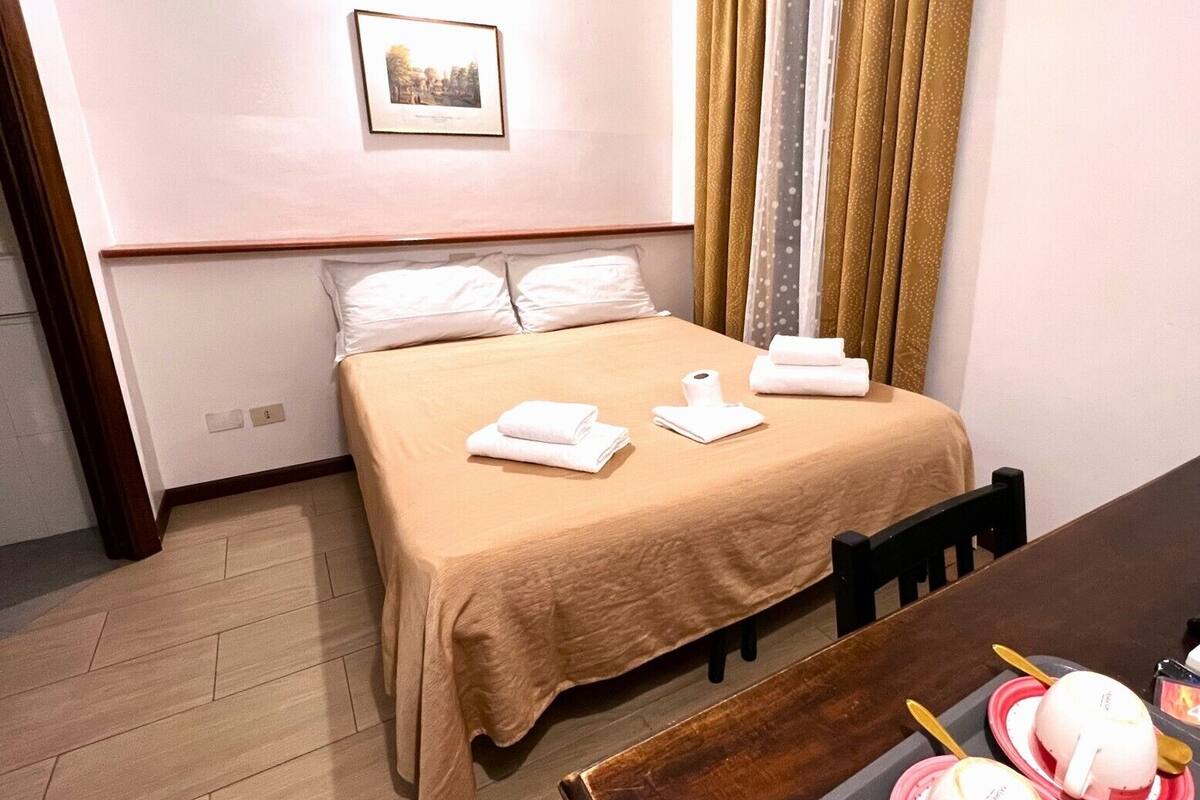 A neatly made bed with white linens, a framed picture on the wall, and a dining table with a pink bowl and utensils.
