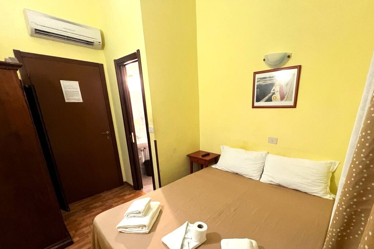 A hotel room with a bed, a nightstand, a picture on the wall, and an air conditioning unit.