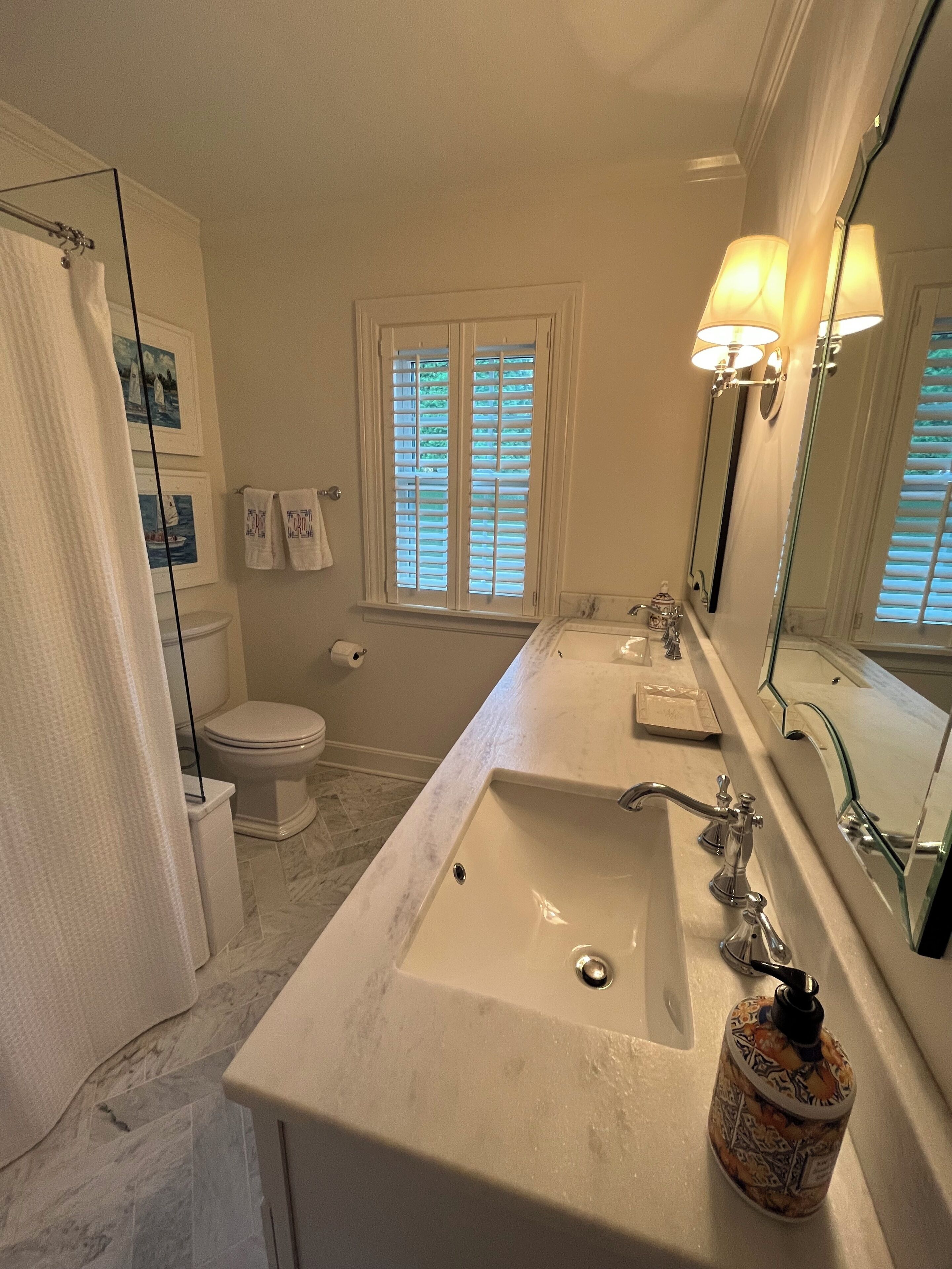 A bathroom with a large sink, two mirrors, a toilet, and a window with shutters.