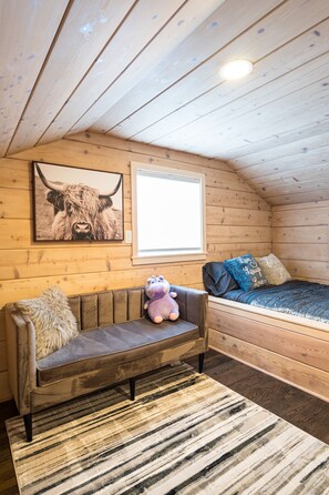 Ett mysigt rum med ett träpanelat tak, en soffa med en fäll, en säng med blå och beige kuddar och en inramad tavla som föreställer en tjur.