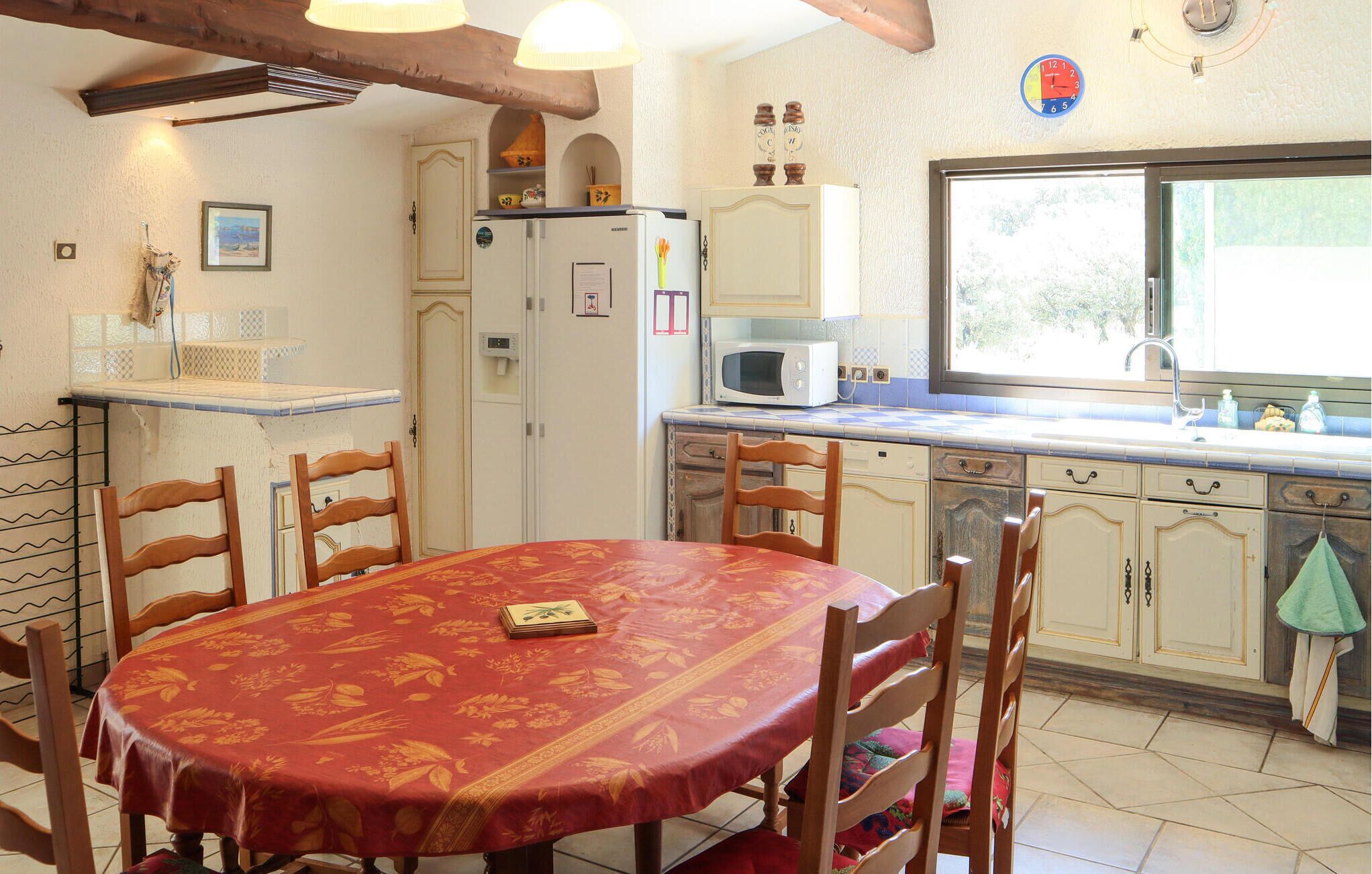 Un coin repas avec une table recouverte d’une nappe rouge, des chaises en bois, un réfrigérateur blanc, un four à micro-ondes et une fenêtre donnant sur un paysage verdoyant.