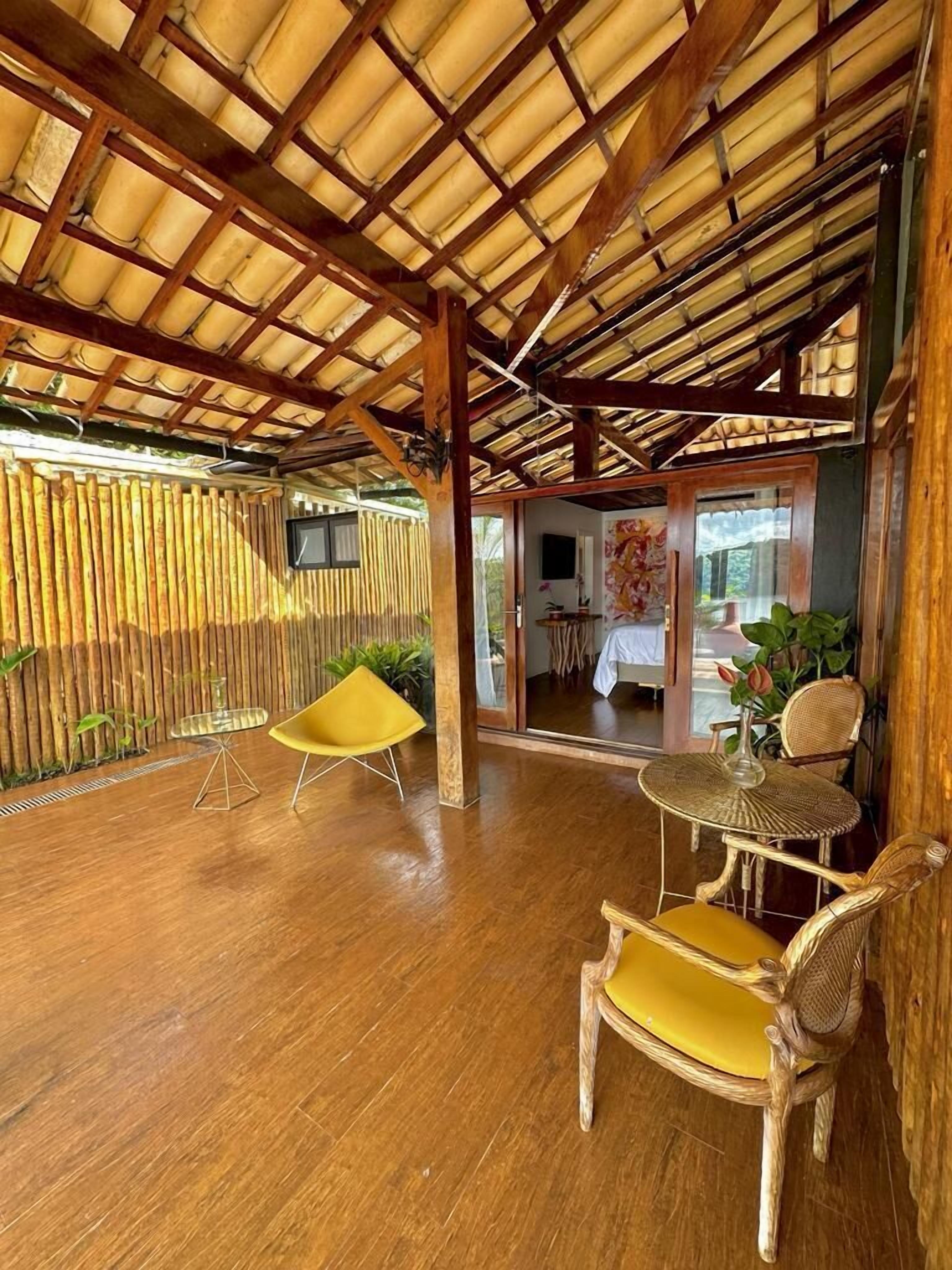 A spacious room with a wooden floor, a bamboo wall, and a ceiling with wooden beams. There is a yellow chair and a wicker table with a vase of flowers.