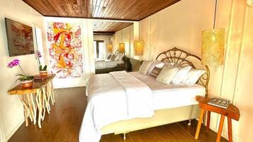 Un dormitorio con techo de madera, cama con ropa blanca, una mesita con lámpara y una pared decorada con un cuadro floral.
