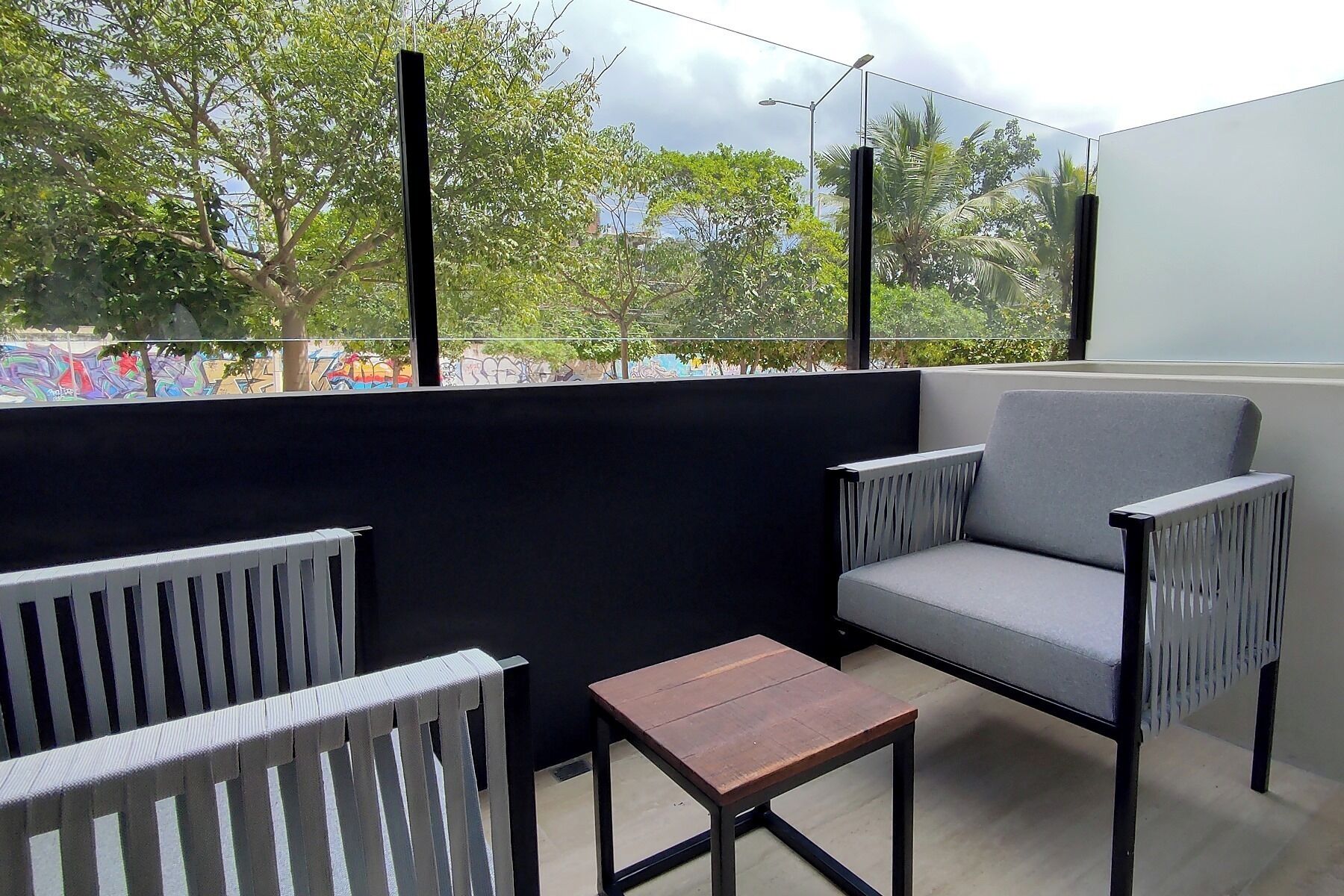 Ein Balkon mit schwarzer Wand, zwei grauen Stühlen, einem kleinen Holztisch und Blick auf Bäume und Graffiti.