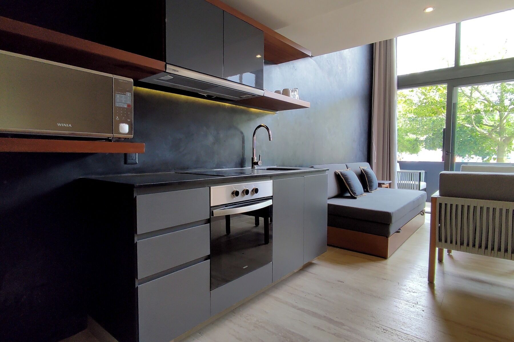 A modern kitchen with a built-in oven, a sink, and a stove. There is a window providing natural light and a view of the outdoors.