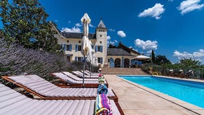 A poolside area with sun loungers, a pool, and a large building with a tower in the background.