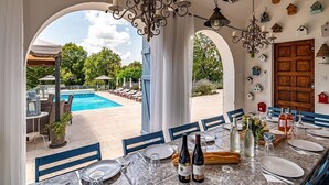 An outdoor dining area with a view of a pool and garden, featuring a table set for dining, a chandelier, and birdhouse decorations on the walls.
