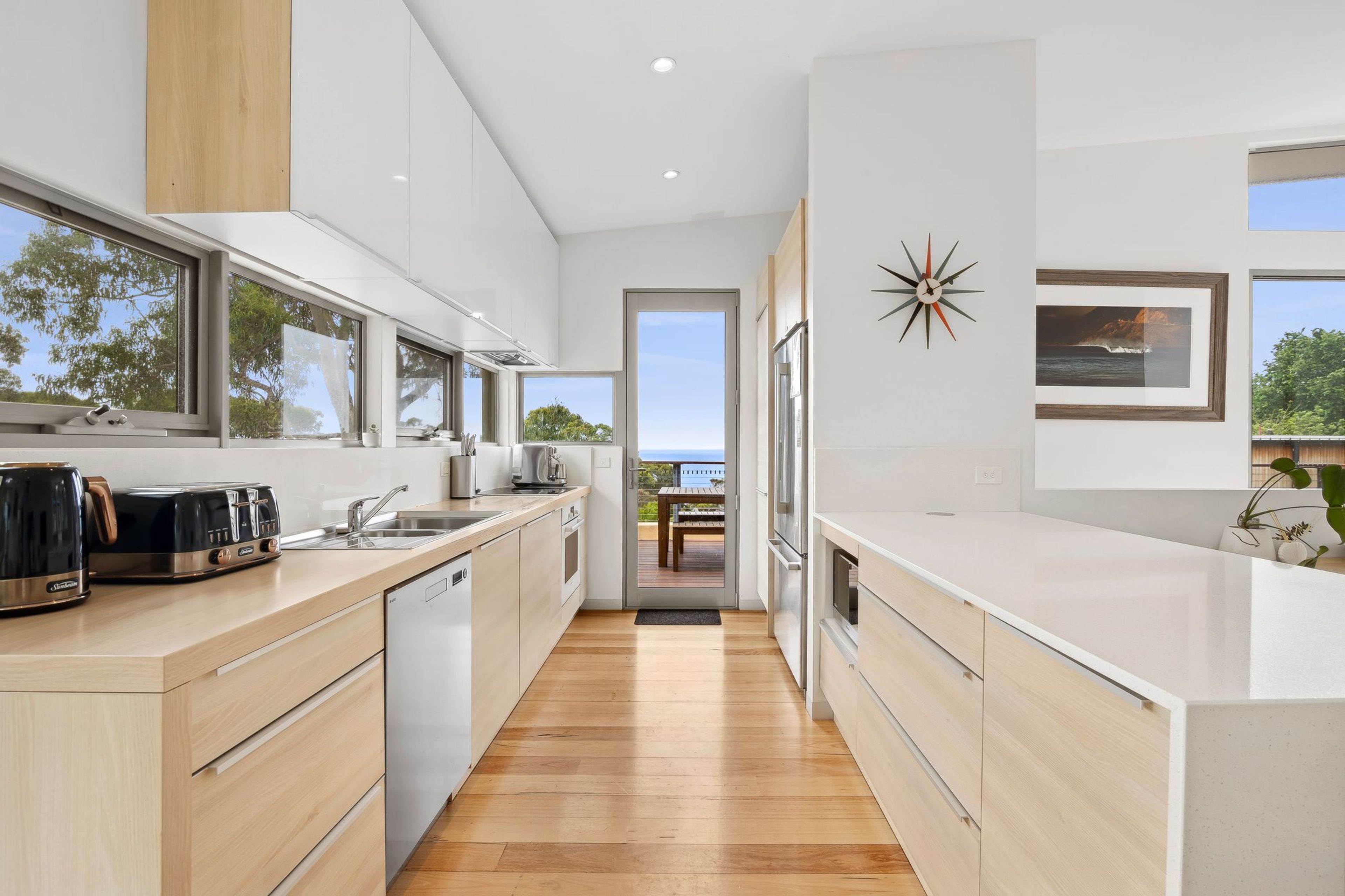 A modern kitchen with wooden flooring, white cabinetry, and large windows offering a view of the outdoors.