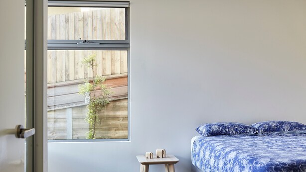 A bedroom with a bed, a small wooden stool, a window, and a view of a wooden fence and a tree.