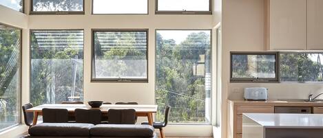 Ein modernes Interieur mit Essbereich, einer Couch, einer Küche und großen Fenstern mit Blick auf Grünflächen.