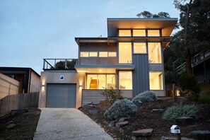A modern two-story house with a visible garage, a covered outdoor area, and a landscaped garden with shrubs and rocks.