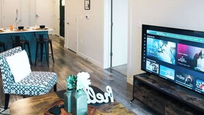 Uma sala de estar moderna com TV exibindo um serviço de streaming, um sofá, uma mesa de centro e uma cozinha ao fundo.