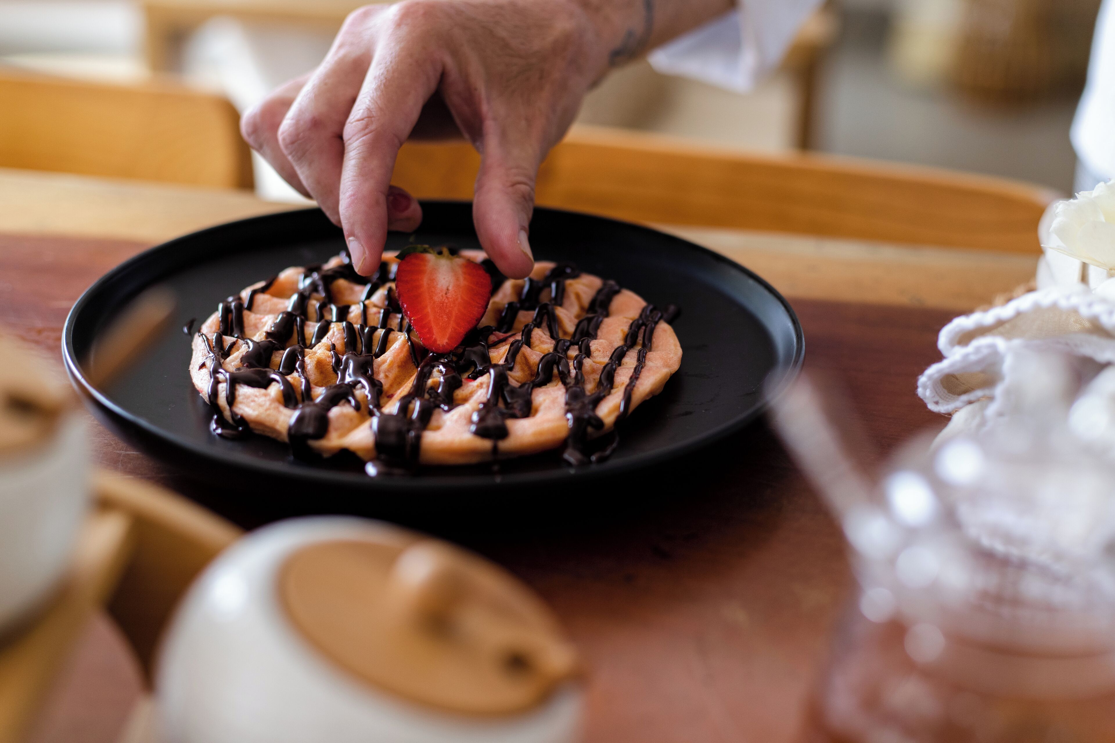 Une main dépose une fraise sur une gaufre enrobée de chocolat, le tout présenté sur une assiette noire.