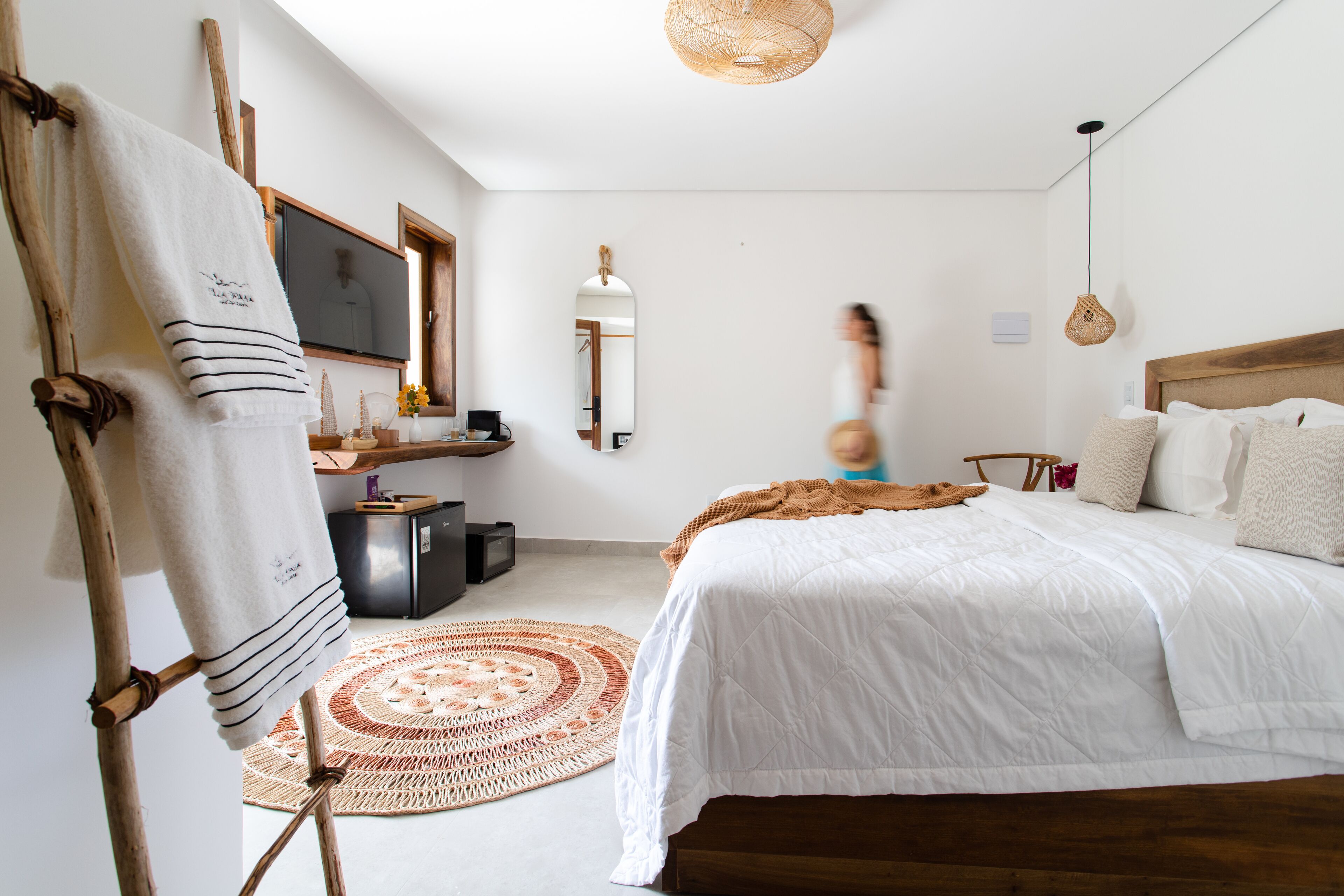 Un dormitorio con una cama, cabecera de madera, alfombra tejida, una silla de madera, una mesita, un televisor y un espejo.