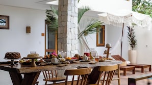 Une table de salle à manger en bois, dressée avec différents plats et boissons, entourée de chaises en bois, dans une pièce dotée d’un pilier en pierre et de grandes fenêtres.