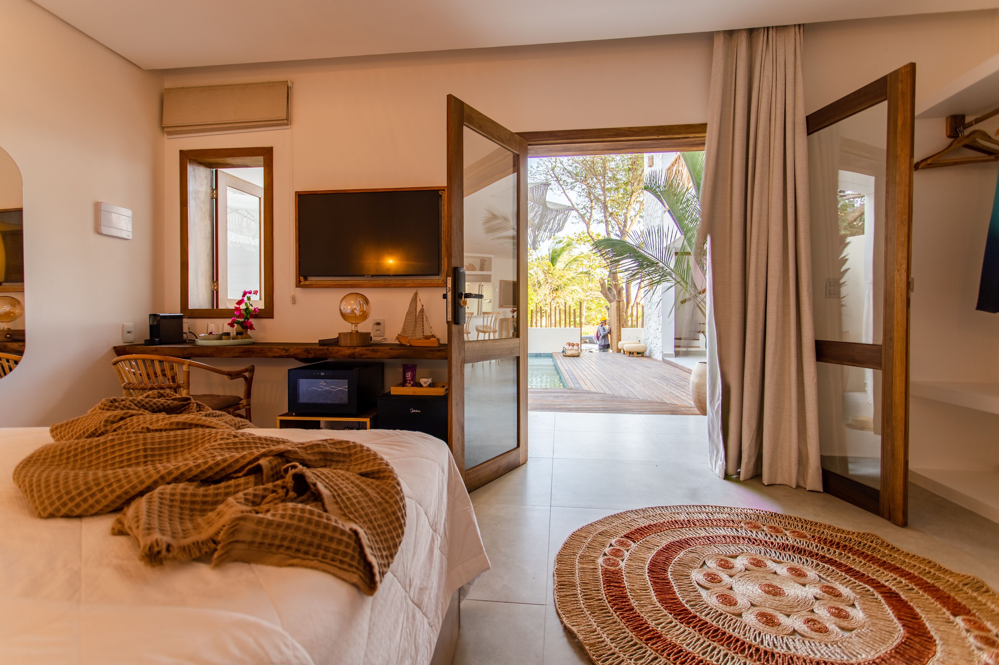 A bedroom with a bed, a desk, a television, and a view of a pool and palm trees through an open door.
