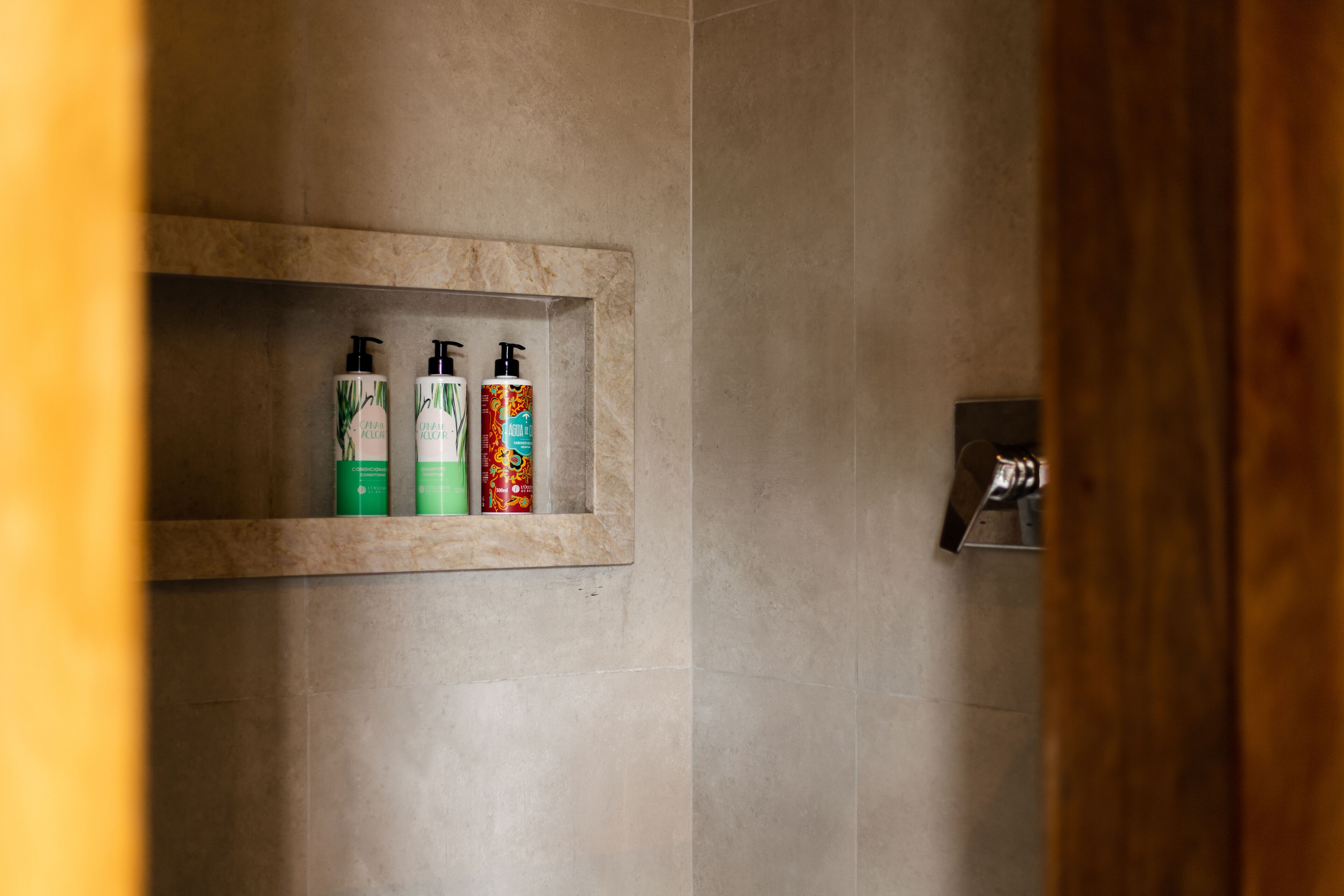 Un baño con pared de mármol, un estante con tres artículos de aseo y un cabezal de ducha.