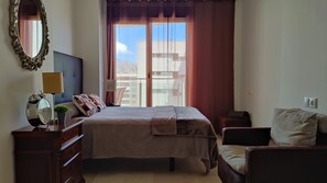 A bedroom with a bed, bedside tables, a lamp, a mirror, and a view of buildings through the window. - CALA ALTA sunrise apartments (Benidorm)