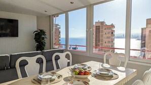 A dining area with a table set for four, a large window with a view of the sea and buildings, and a plant on the left. - DON MIGUEL blue sky apartment (Benidorm)