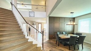 A spacious dining area with a wooden table and chairs, a staircase with a railing, and a clock on the wall.