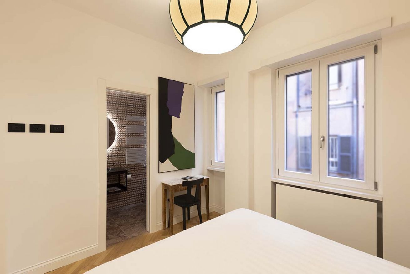 A room with a bed, a desk, a chair, a window, and a door leading to another room.