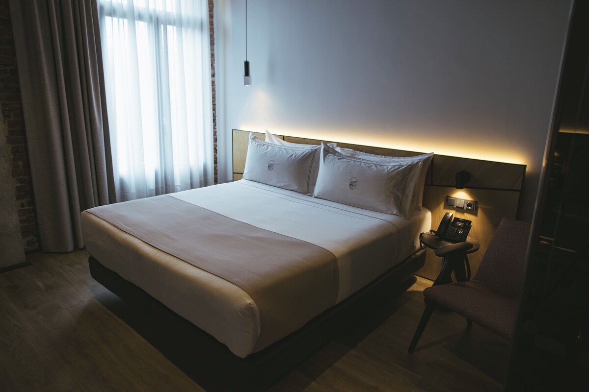 Een netjes opgemaakt bed met een houten hoofdbord, een nachtkastje met een telefoon en een stoel.