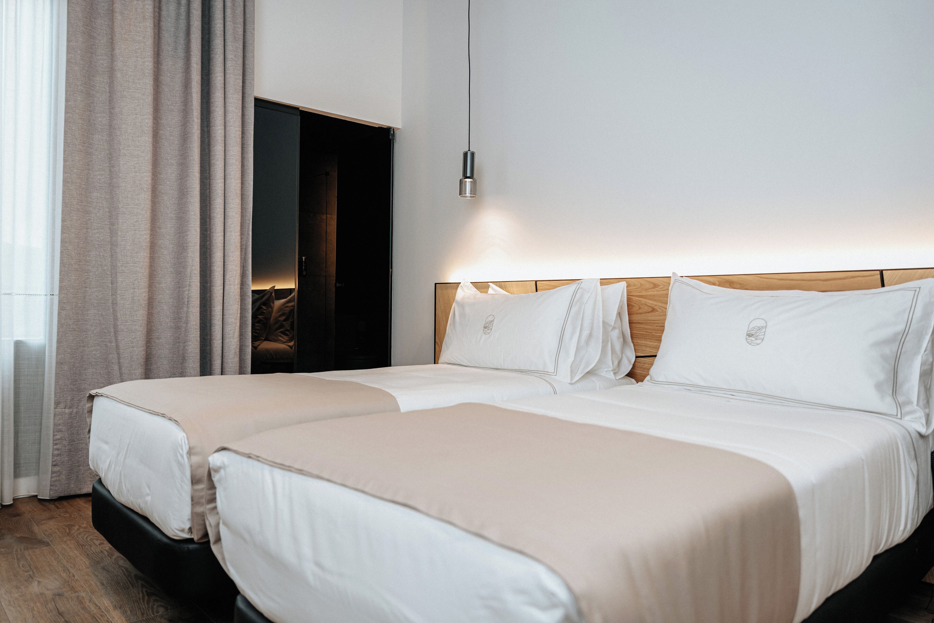 A hotel room with two beds, a wooden headboard, a hanging light fixture, and a view of the outdoors through a window with curtains.