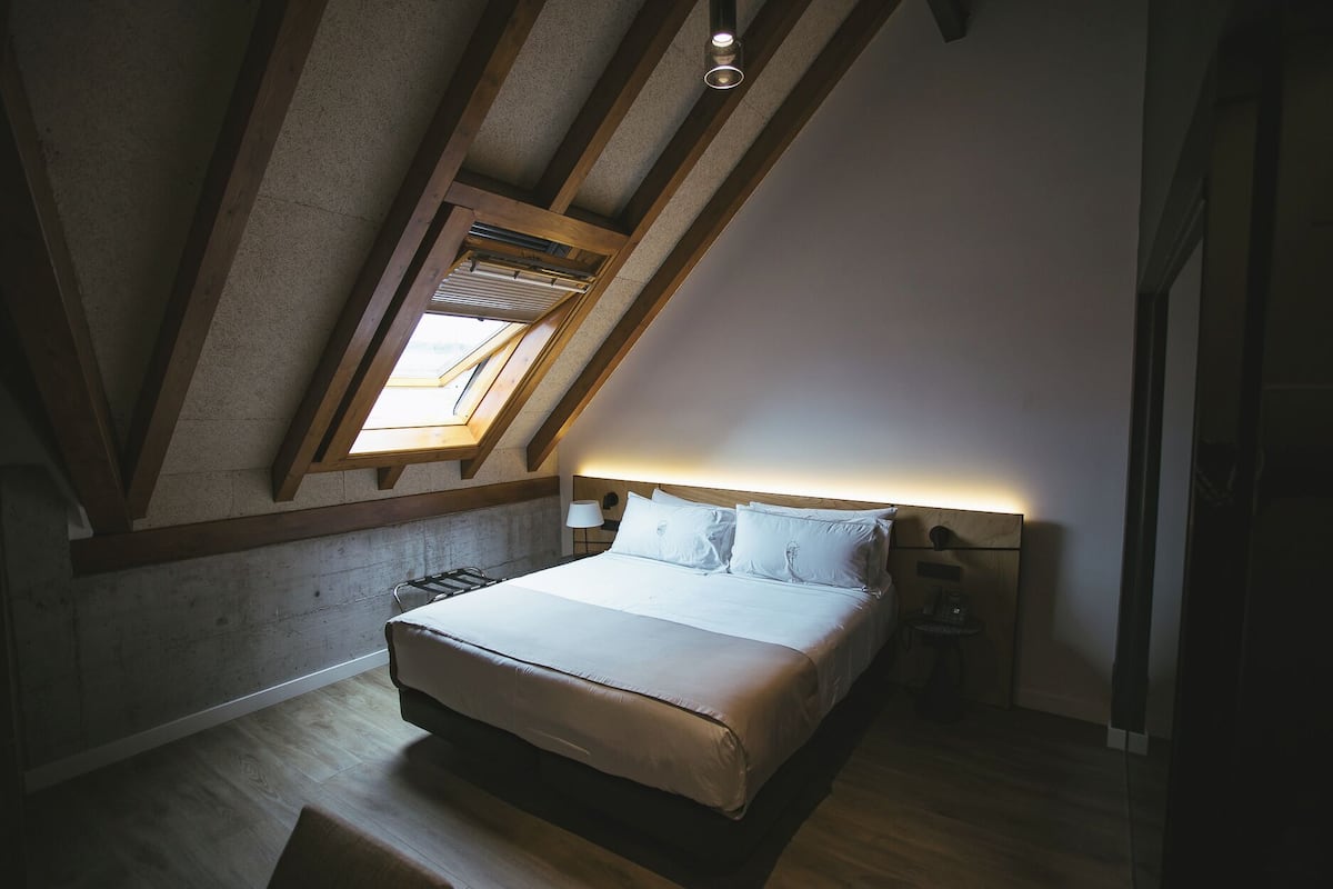 A bedroom with a sloped ceiling, skylight, wooden beams, a bed with white bedding, a bedside table, and a nightstand.