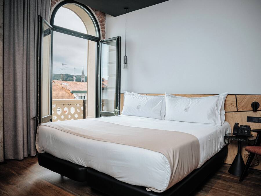 A modern hotel room with a large bed, a wooden headboard, a small bedside table with a phone, and a view of the city through a large window.