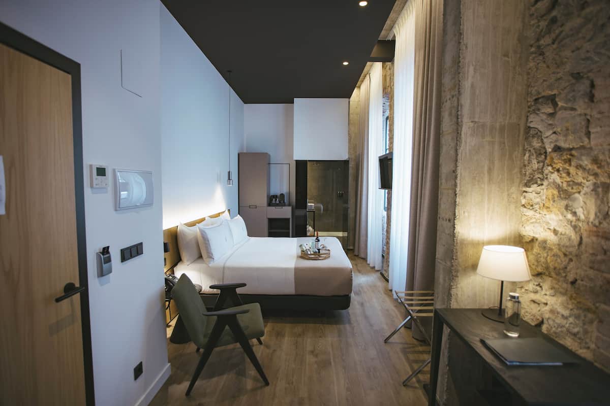 A modern hotel room with a bed, bedside table, chair, and a stone accent wall.