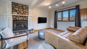 A living room with a stone fireplace, a leather sofa, a white armchair, and a flat-screen TV mounted on the wall. - PRESIDENTIAL DUPLEX SUITE WITH HOT TUB AND WRANP AROUND BALCONY (Windham)