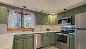 Una cocina con gabinetes verdes, electrodomésticos de acero inoxidable, lavavajillas y ventana con vistas a árboles.
