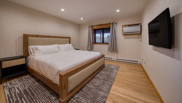 Un dormitorio con una cama grande, una mesita de noche, un televisor montado en la pared y una ventana con cortinas.