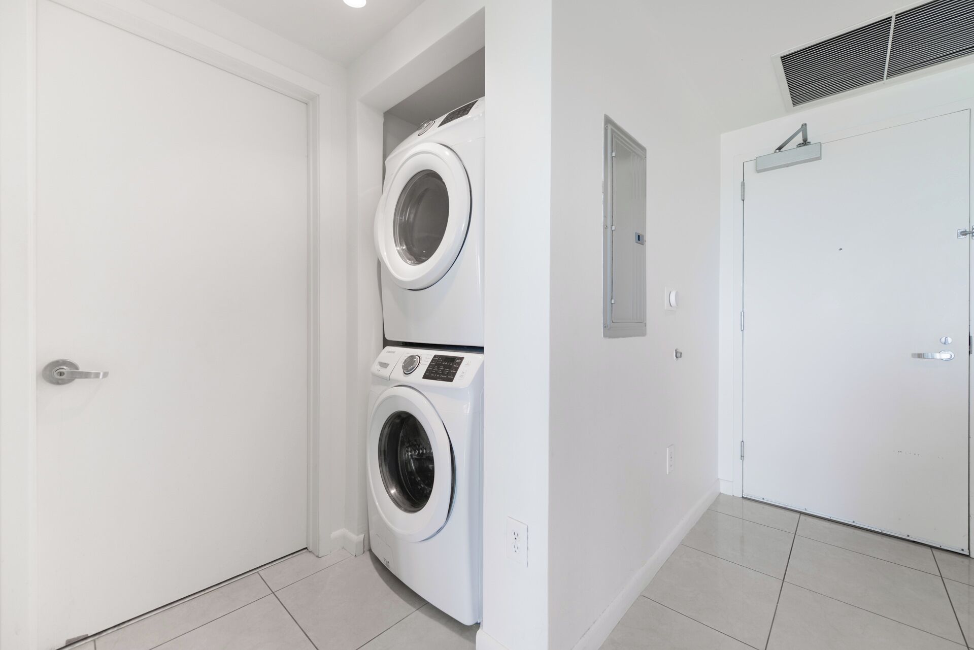 A laundry room with two stacked washing machines, a door, and a vent on the ceiling.