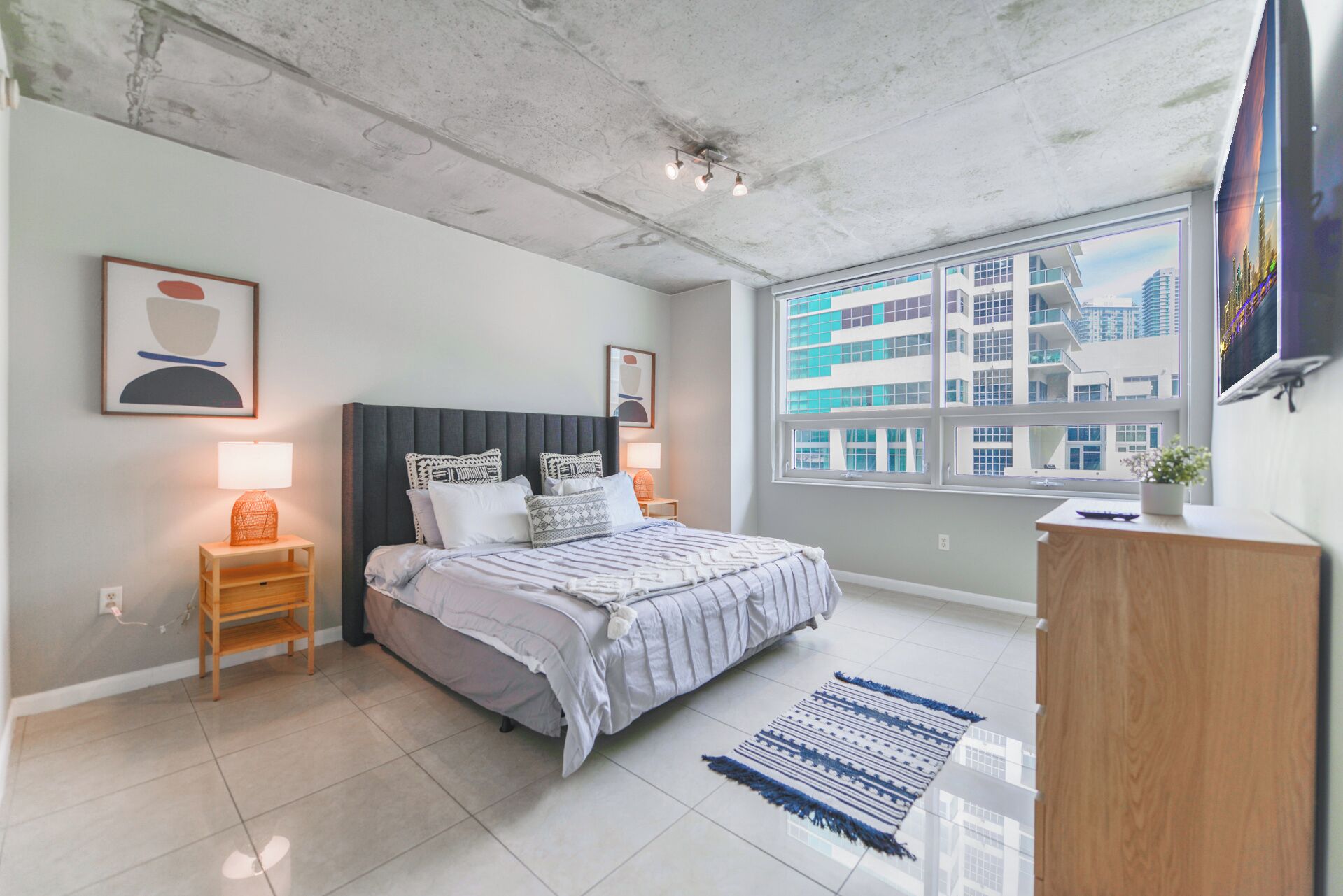 A modern bedroom with a bed, bedside tables, a nightstand, a dresser, a window with a city view, and a ceiling with exposed beams.