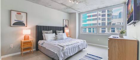 A modern bedroom with a bed, bedside tables, a nightstand, a dresser, a window with a city view, and a ceiling with exposed beams.