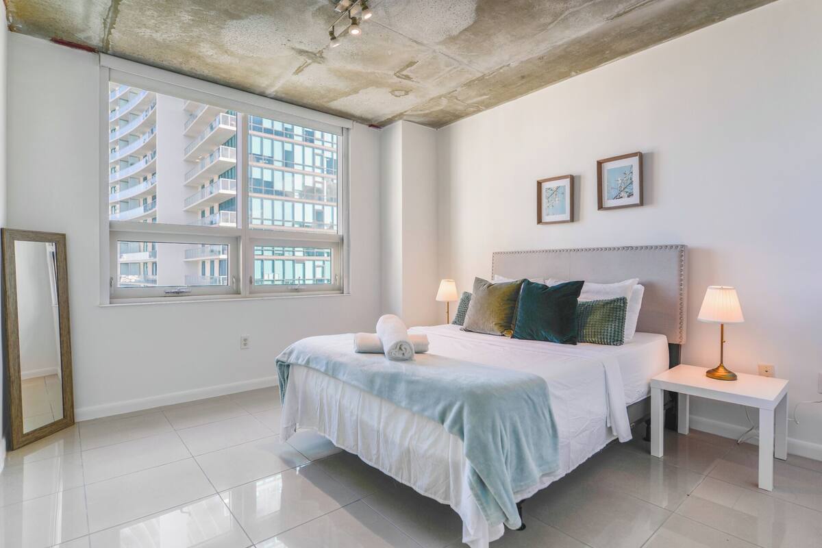 A modern hotel room with a large bed, two bedside tables, a large window with a view of a building, and a ceiling with exposed concrete.