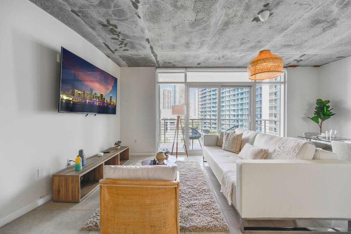 A modern living room with a large TV, a sofa, a coffee table, and a view of high-rise buildings.