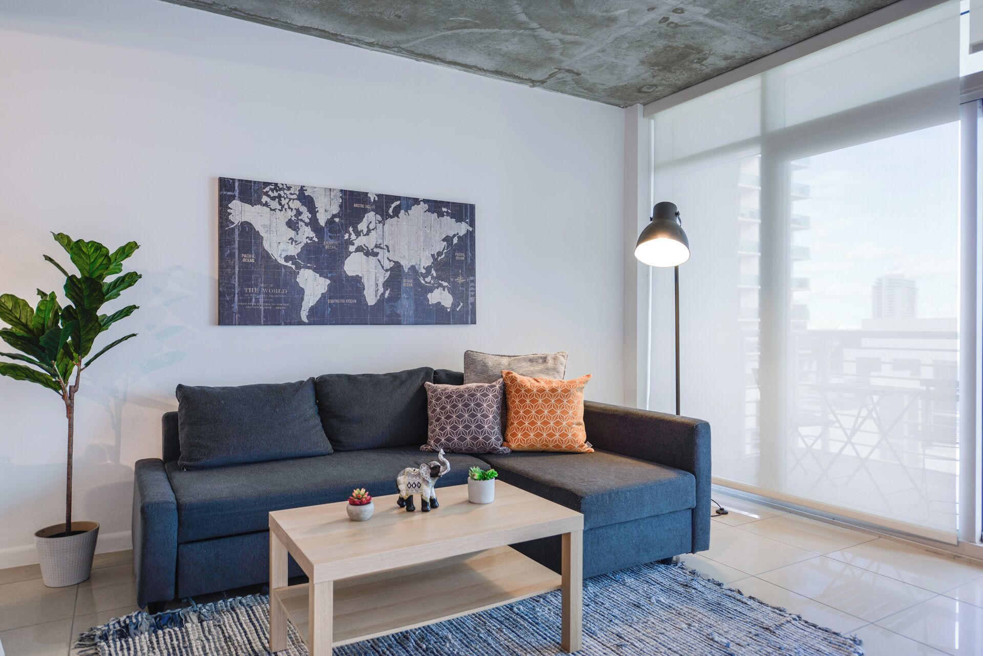 A modern living room with a blue sofa, a world map artwork, a coffee table, and a large window with a city view.